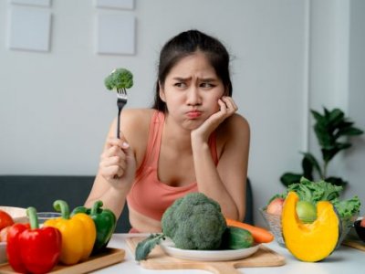 An unimpressed woman holding up a piece of broccoli on a fork.